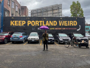 Mann mit Regenschirm vor Schild mit der Aufschrift Keep Portland Weird