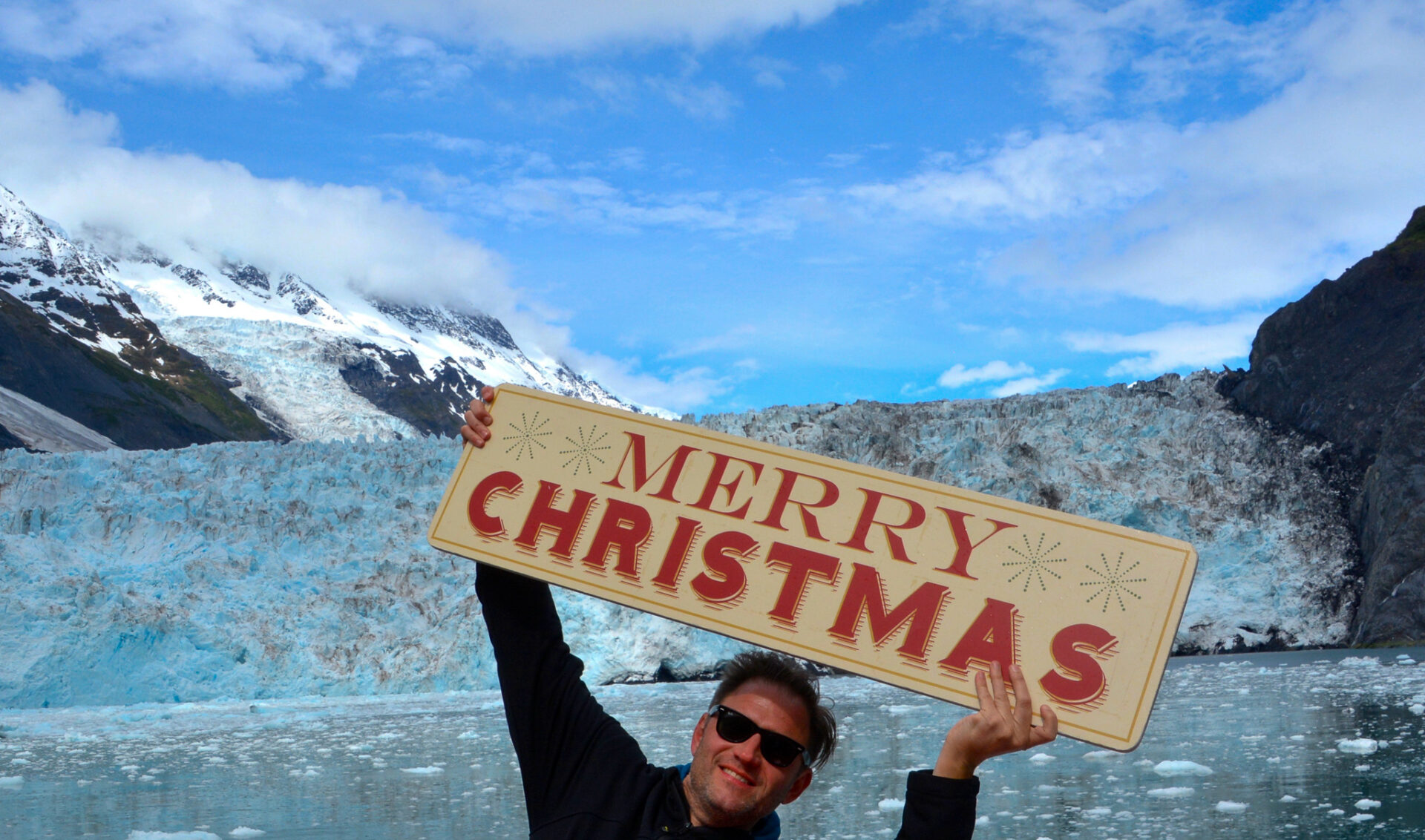 Autor Ralf Johnen hält an Bord der MS Klondike ein Schild mit der Aufschrift Merry Christmas. Im Hintergrund einer der 26 Gletscher der Bootstour